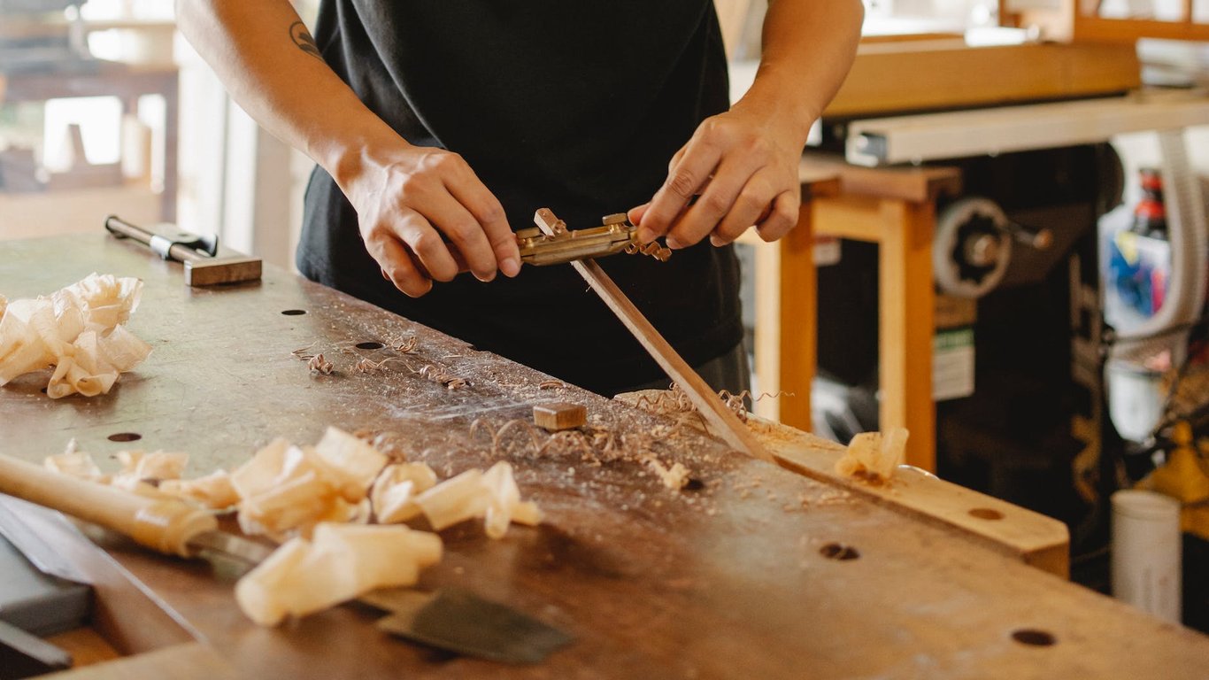 Menuisier anonyme en train de lisser un bâton de bois avec une lame de rasoir, debout à l'établi, dans un atelier, par une journée ensoleillée.